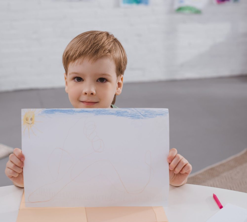portrait of cute boy showing picture in hands at table in classroom